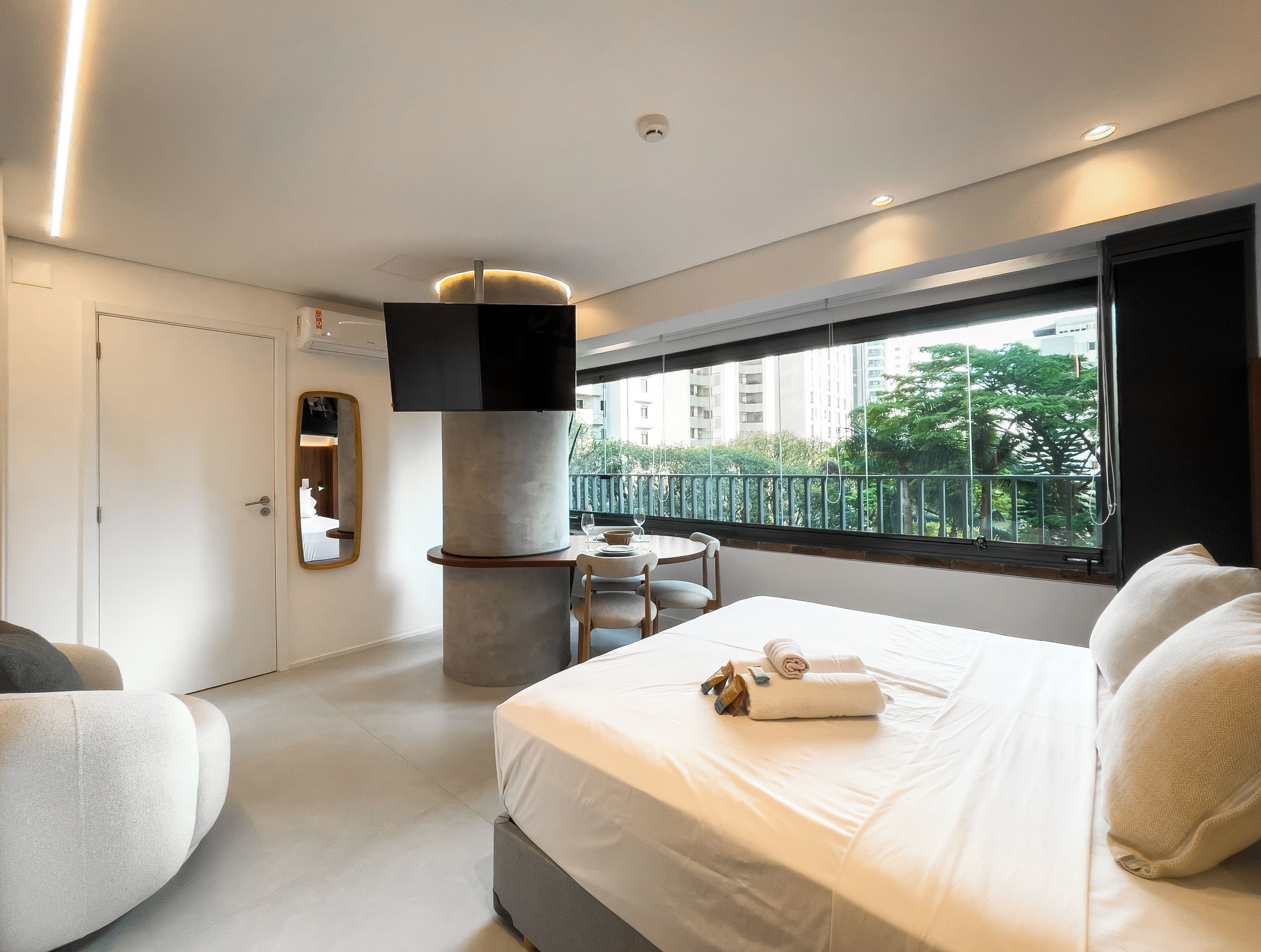 A modern hotel room with a bed, a seating area, a TV, and a balcony with a view of buildings and trees.