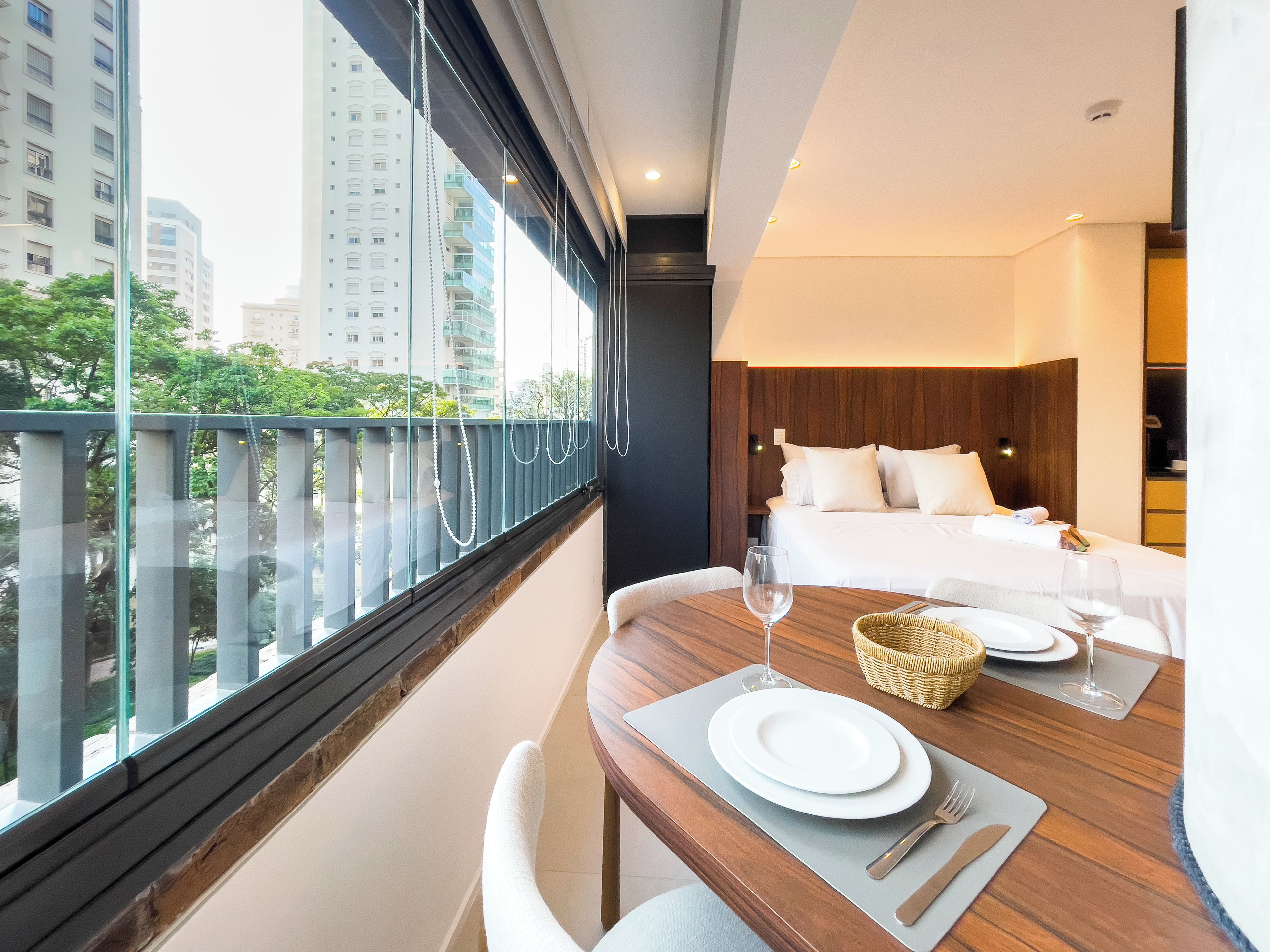 A hotel room with a bed, a dining table set for two, and a view of the city through large windows.