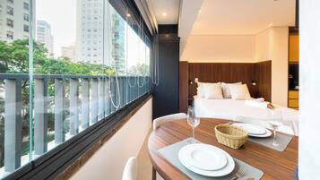A hotel room with a bed, a dining table set for two, and a view of the city through large windows.