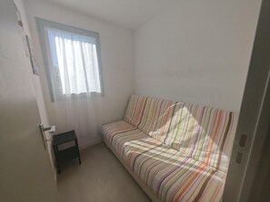 A small, sparsely furnished room with a single striped sofa, a small table, and a window with sheer curtains. - Air-conditioned apartment near beach (Saint-Cyprien)