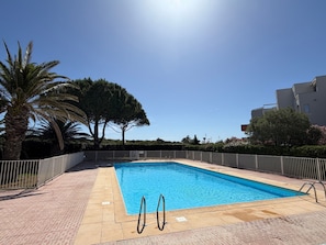 A rectangular swimming pool with a tiled deck, surrounded by a white fence, palm trees, and a building in the background. - Air-conditioned apartment near beach (Saint-Cyprien)