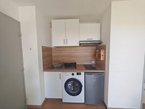A compact kitchen with a washing machine, a small refrigerator, and wooden cabinets. - Air-conditioned apartment near beach (Saint-Cyprien)