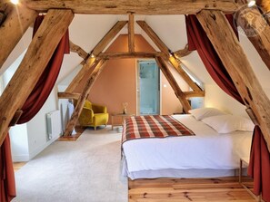 Ein Schlafzimmer mit schrägverlaufender Decke, Holzbalken, einem Bett mit weißen Leinen, einem gelben Sessel und einer rot-weiß karierten Decke.