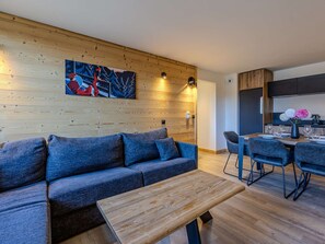 A modern living space with a wooden wall, a sofa, a dining table with chairs, and a kitchen area. - Practical apartment at the ski slopes - 6 guests (Belle Plagne)