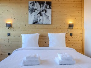 Habitació d'hotel amb un capçal de llit de fusta, dos coixins blancs, un llit amb roba de llit blanca, dues làmpades fixes a la paret i un quadre amb pingüins.