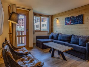 A cozy living room with a sofa, a coffee table, and a leather chair. There are windows with curtains, a painting on the wall, and a wooden floor. - Practical apartment at the ski slopes - 6 guests (Belle Plagne)