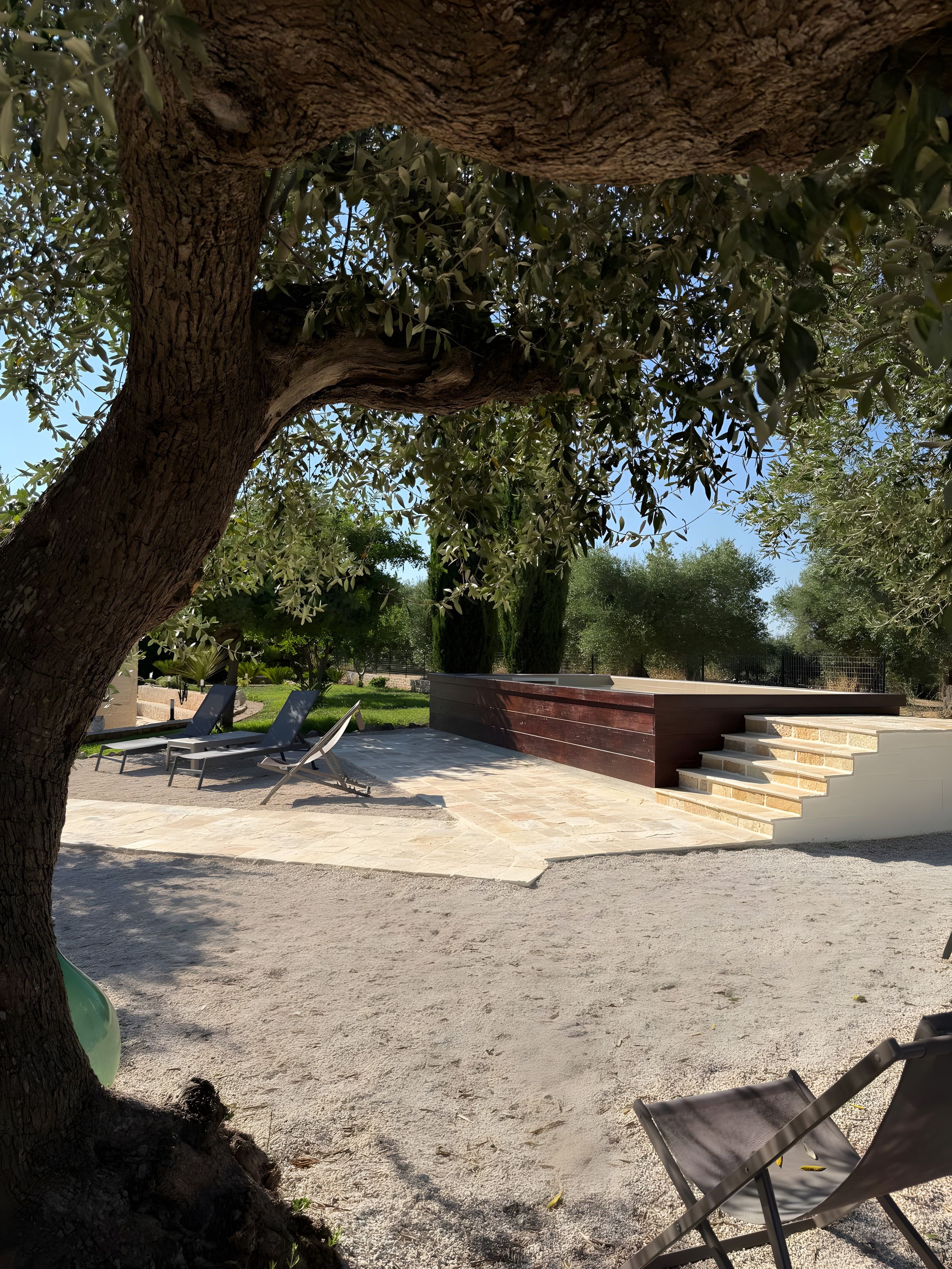 A patio area with a tree providing shade, a set of stairs leading to a higher level, and a few lounge chairs.