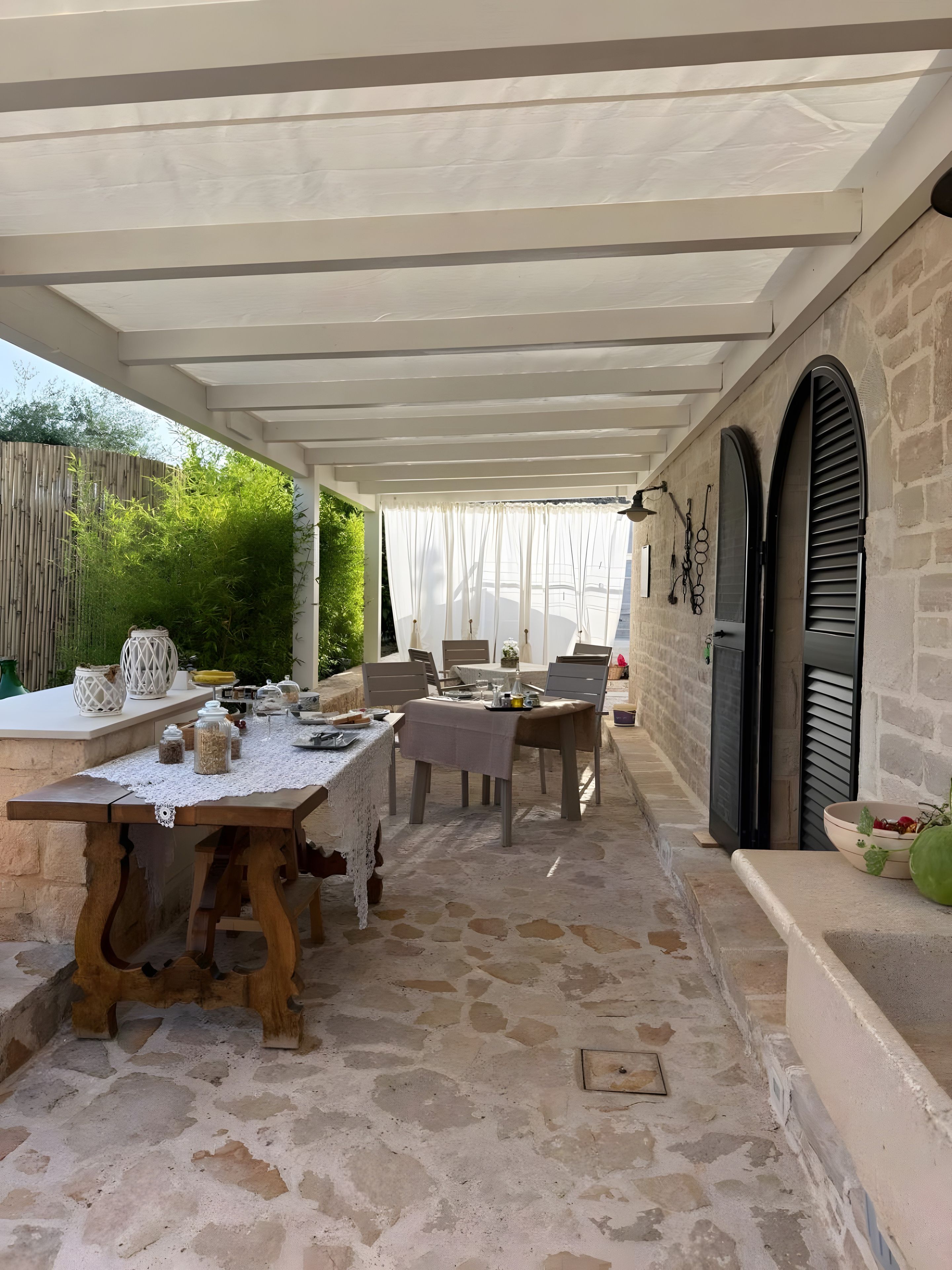 Une terrasse extérieure avec une table dressée pour les repas, un sol en pierre et un espace couvert doté d’un plafond blanc.