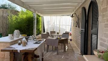 Une terrasse extérieure avec une table dressée pour les repas, un sol en pierre et un espace couvert doté d’un plafond blanc.