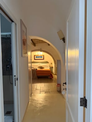Un pasillo que conduce a un dormitorio con una cama, un cuadro en la pared y una zona de ducha con azulejos.