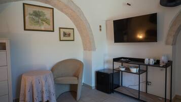 A room with a TV mounted on the wall, a small table with a tablecloth, a chair, and a cabinet with shelves.
