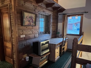 A wooden cabin interior with a television, a window with bars, a picture on the wall, and a view of a building outside. - Typical Chalet 6 Rooms, Garden, 8 Pers (Saint-Martin-de-Belleville)