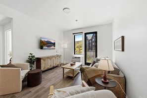 A modern living room with a sofa, armchair, coffee table, and a dining table. There is a large window with a view of a building and a landscape. - Pioche Basecamp | Deer Valley + Mountain Views (Park City)