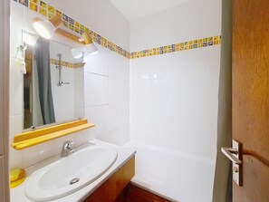 Ein Badezimmer mit einem weißen Waschbecken, einem Spiegel und einer Dusche. Über dem Spiegel befinden sich zwei Lampen und ein Handtuchhalter.