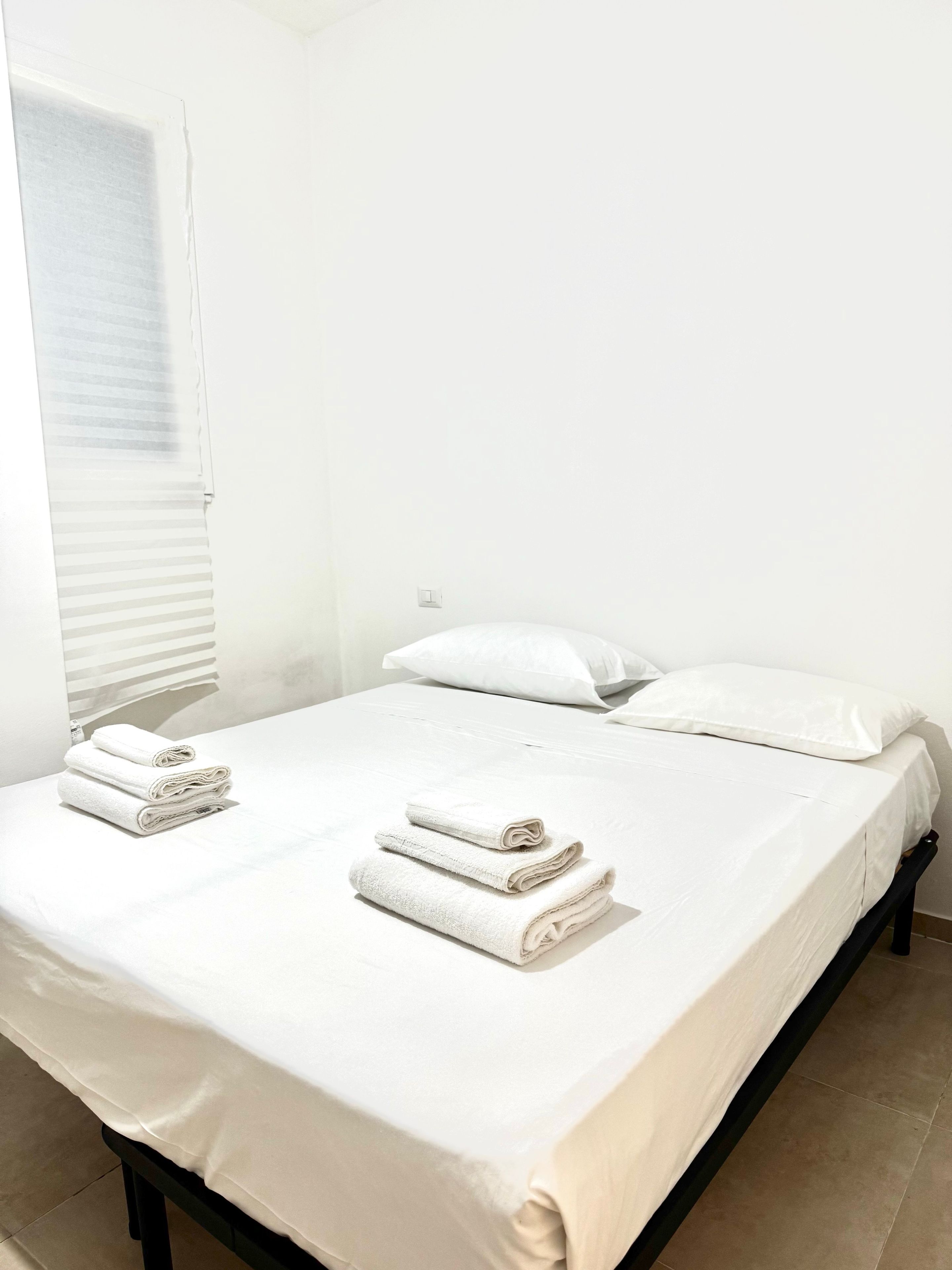 Un lit simple avec du linge de lit blanc et des serviettes pliées, une fenêtre avec des stores et un mur sobre.