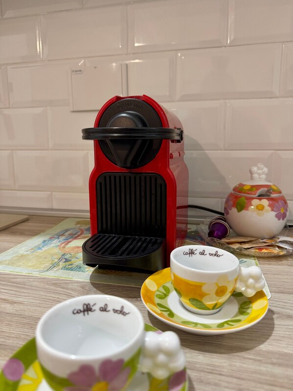 Una máquina de espresso roja sobre una encimera de cocina, con una taza y platillo de café, un elegante hervidor y un posavasos.