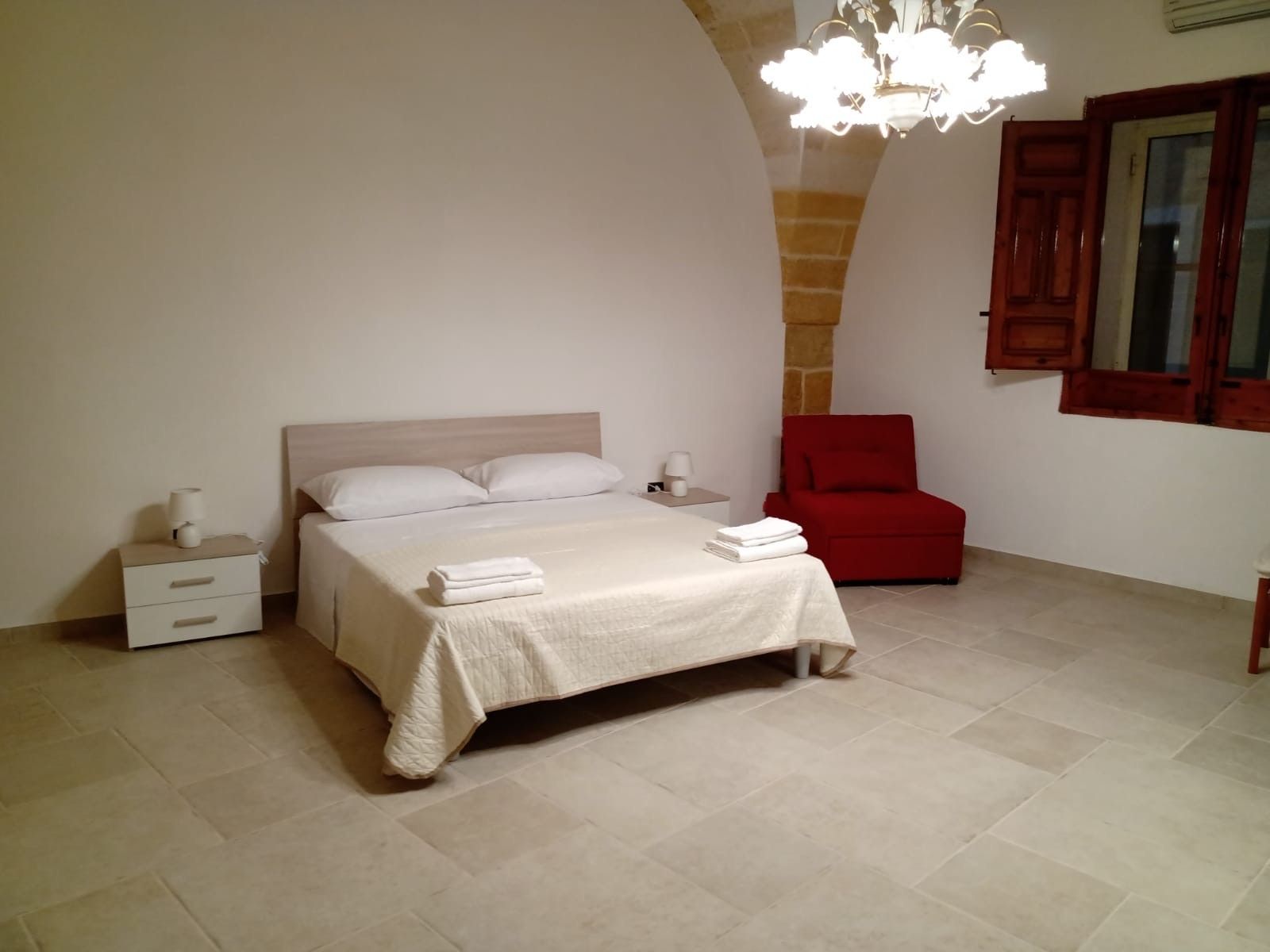 A bedroom with a bed, nightstands, a red chair, and a chandelier.
