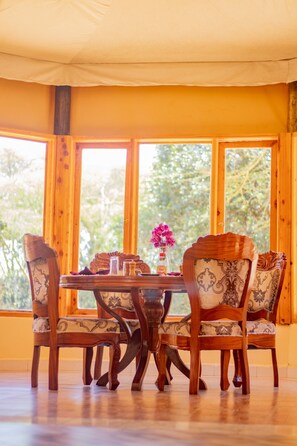 A dining area with a wooden table and chairs, a vase with flowers, and a bottle of sauce. - Nyota Springs Luxury Camp (Maasai Mara)
