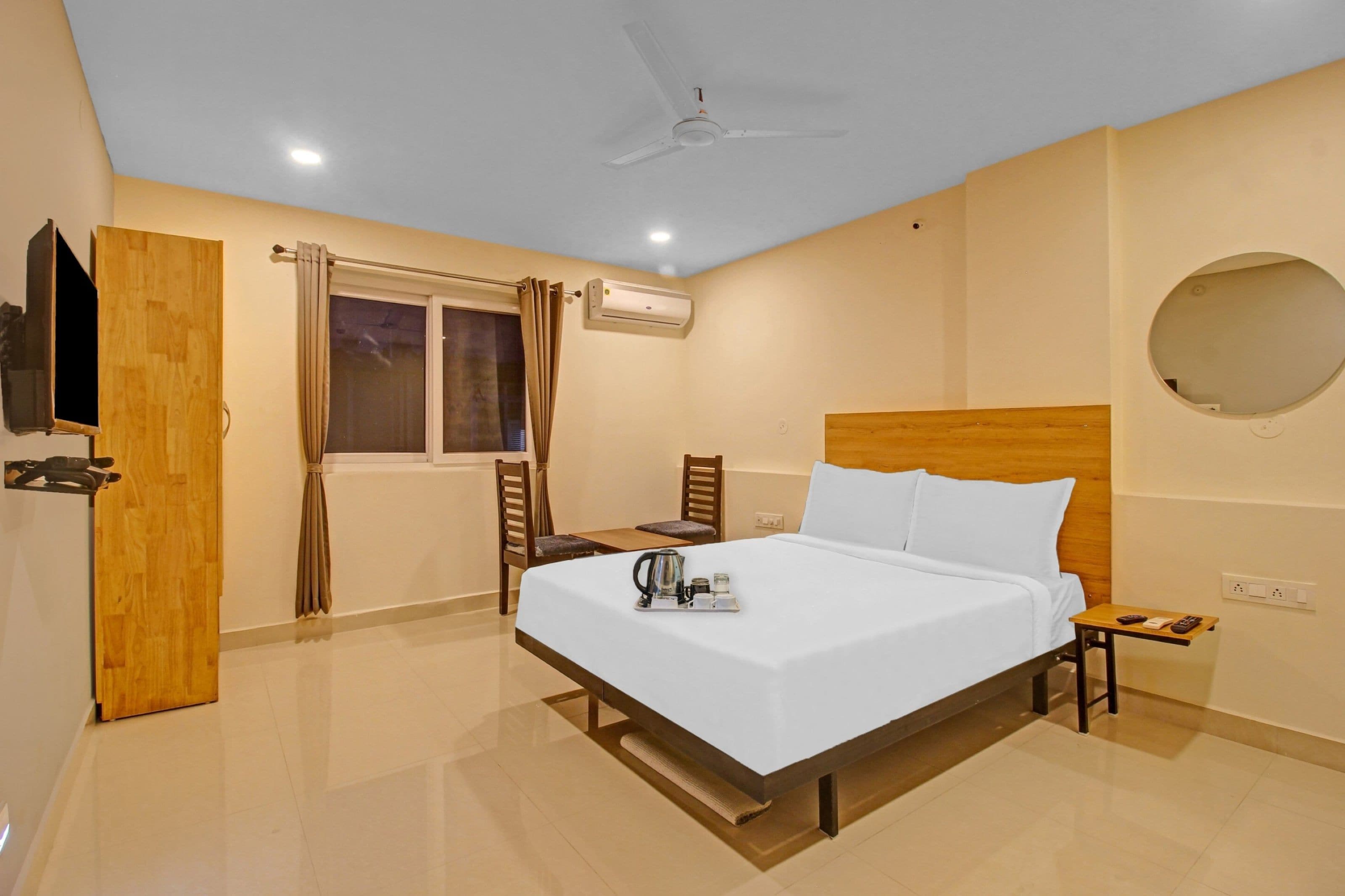 A hotel room with a bed, a bedside table, a chair, a window with curtains, an air conditioning unit, a ceiling fan, and a television mounted on the wall.