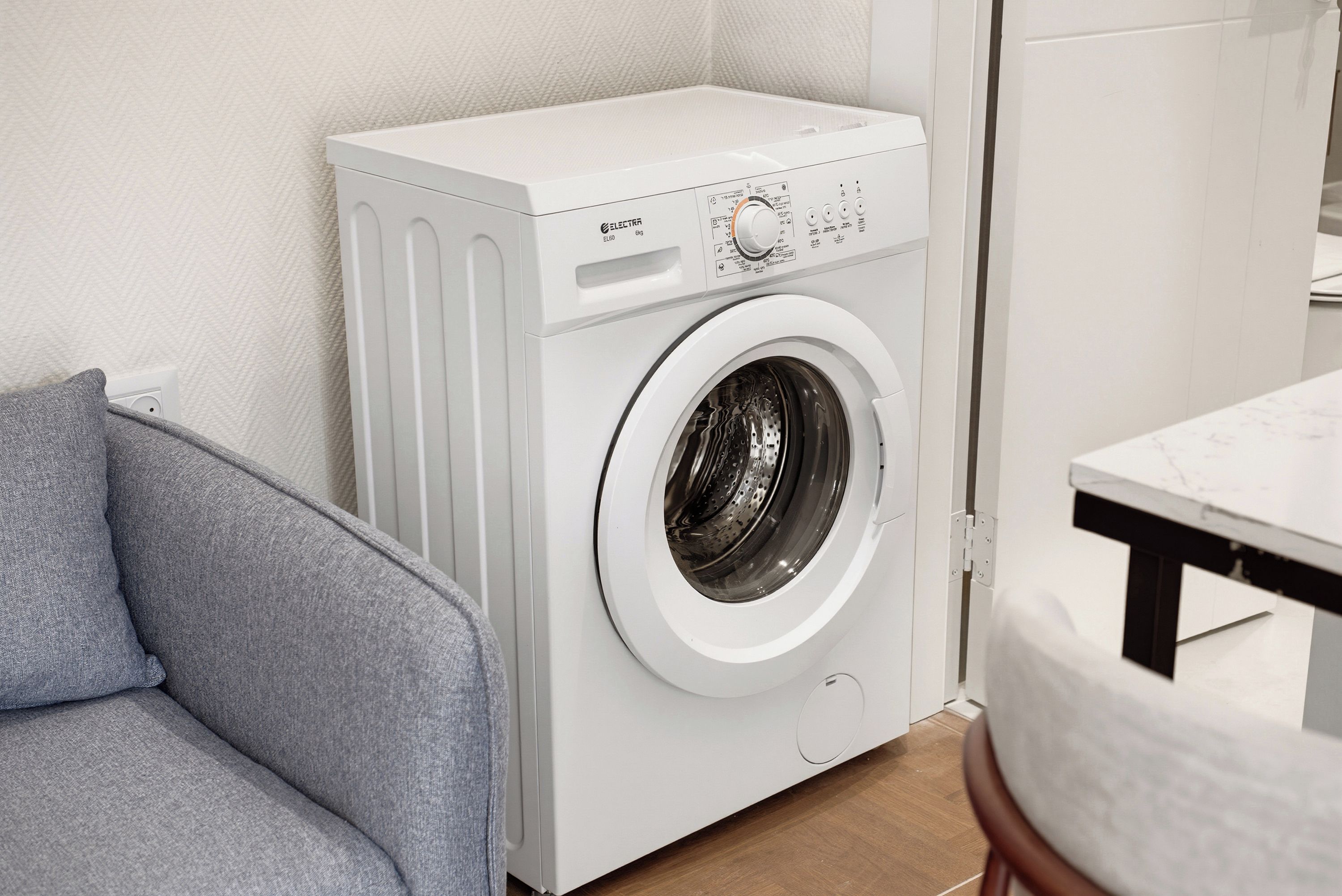 Una lavadora de carga frontal en un cuarto de lavado, con un sofá gris y una mesa de madera.