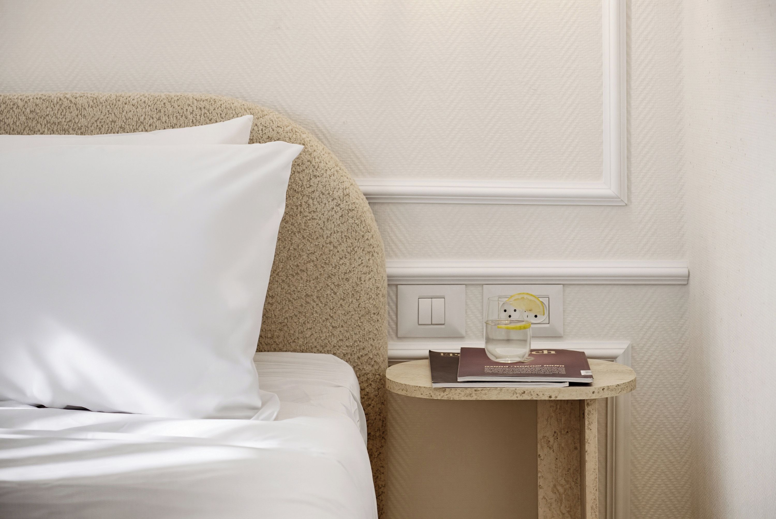 A neatly made bed with white linens and a single pillow, a small bedside table with a book and a glass of water, and a wall switch.