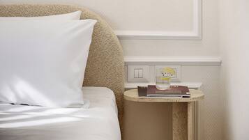 A neatly made bed with white linens and a single pillow, a small bedside table with a book and a glass of water, and a wall switch.