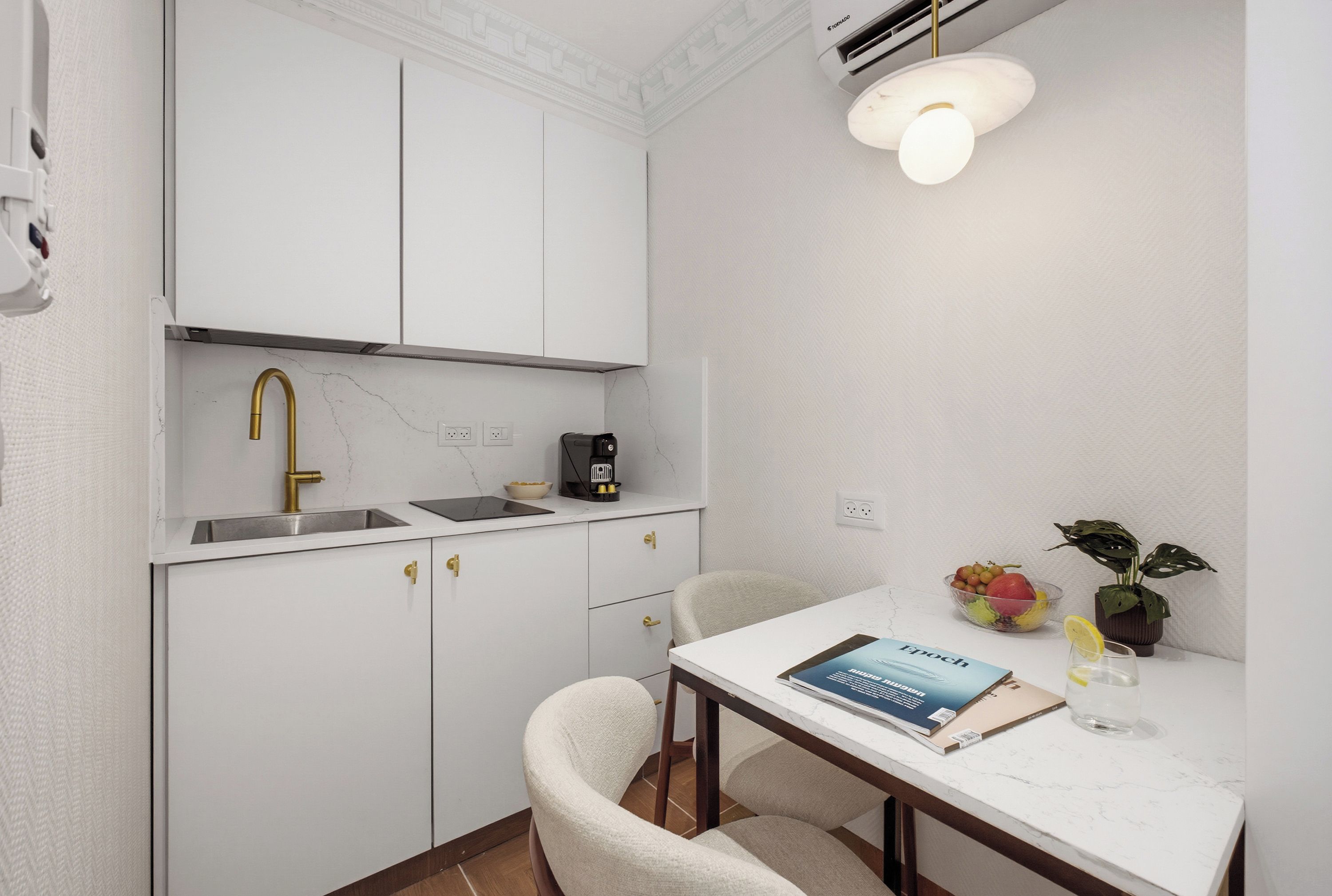 A compact kitchen with a white countertop, a gold faucet, a coffee maker, and a dining area with a chair and a table.