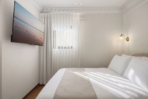 Habitación de hotel con una cama grande, un televisor de pantalla plana montado en la pared, una ventana con cortinas translúcidas y una lámpara de pared.