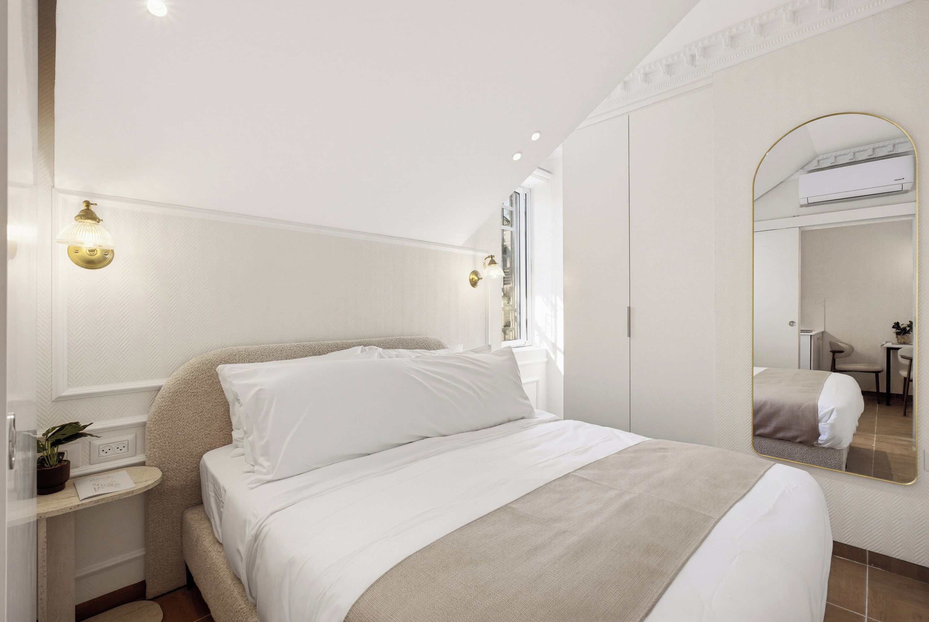 A hotel room with a bed, bedside tables, a mirror, and an air conditioning unit.
