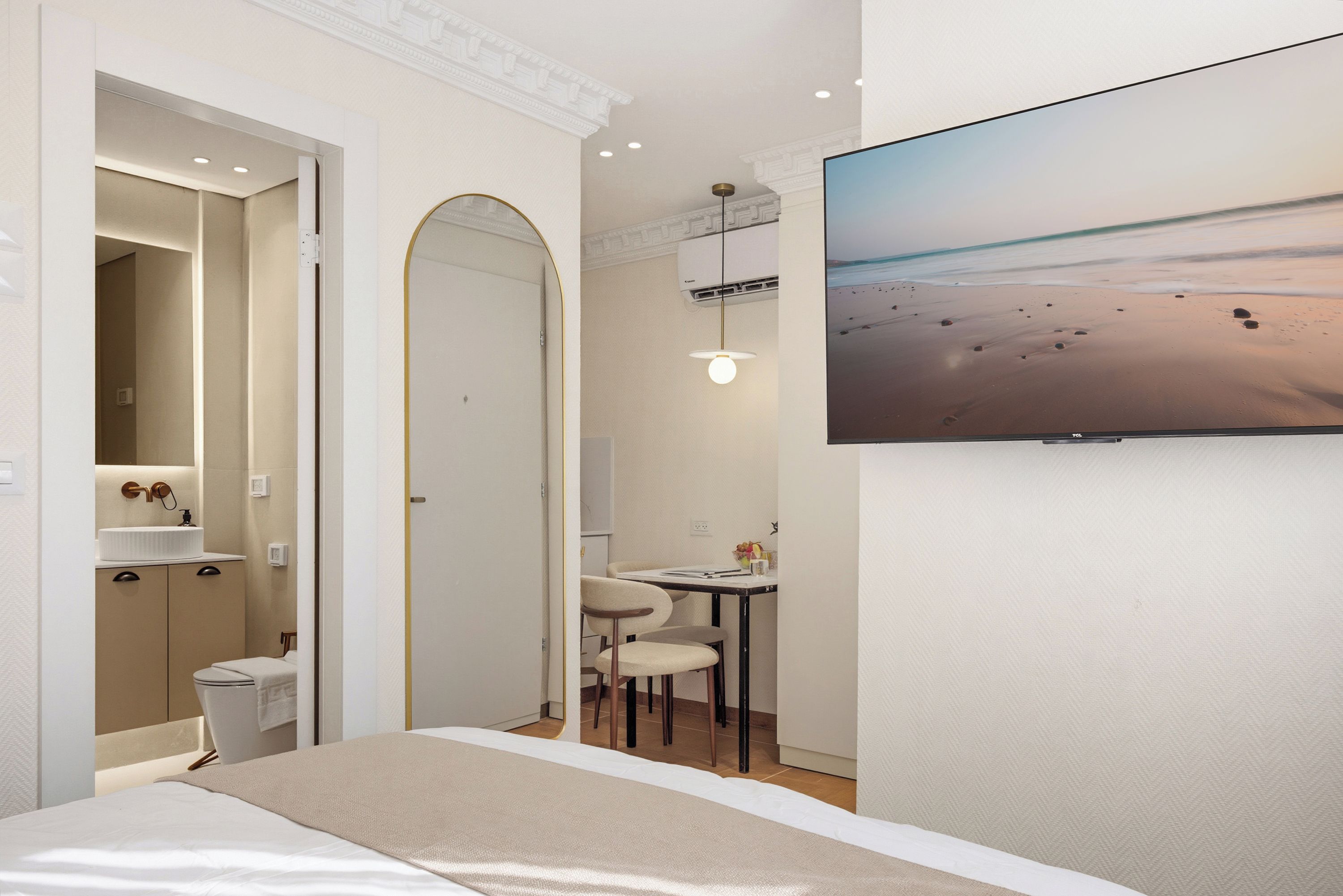 Habitación de hotel con cama, escritorio, silla, televisor montado en la pared y baño con lavamanos y espejo.