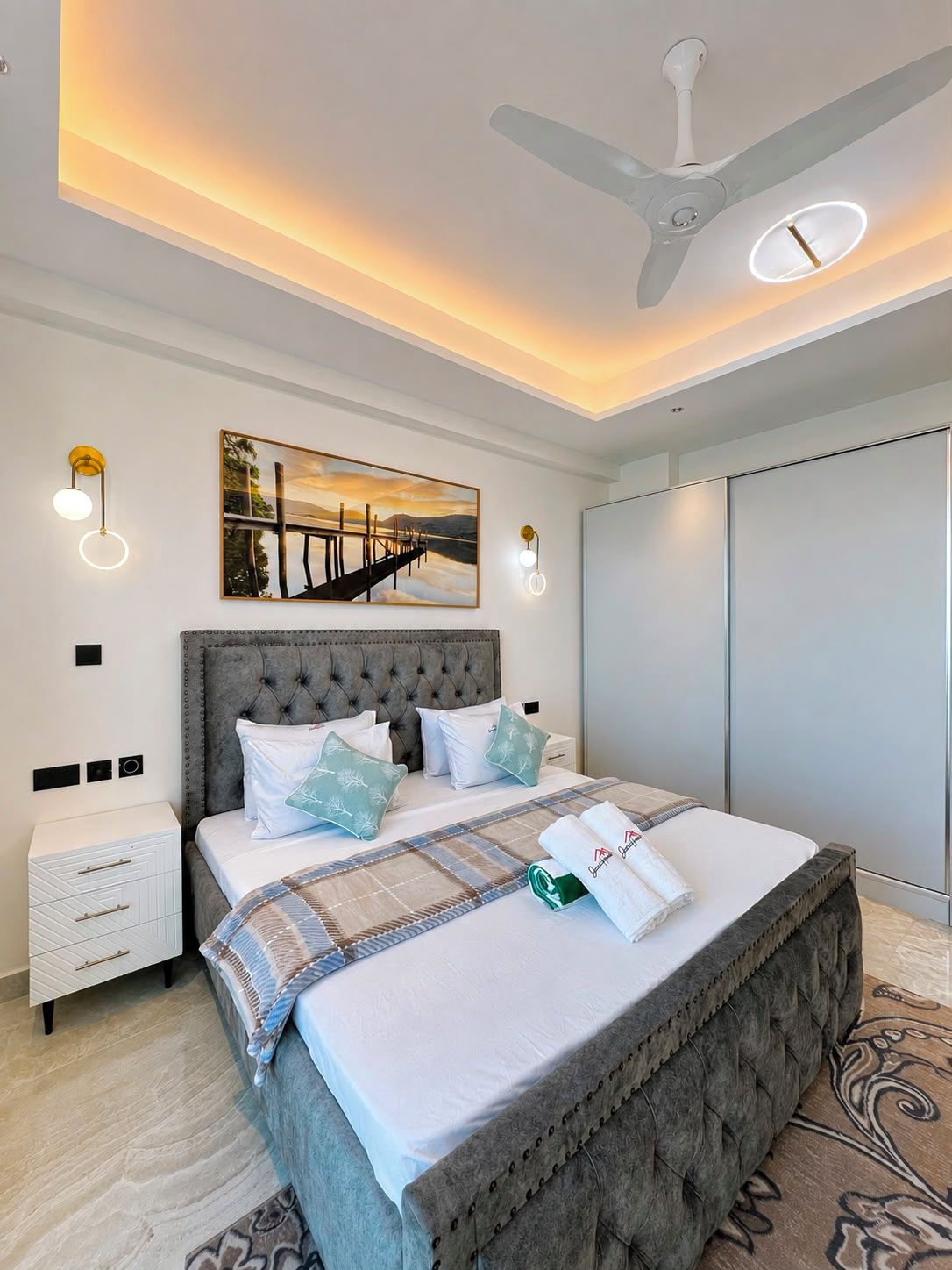 Un dormitorio con una cama grande, un ventilador de techo, luces fijadas en la pared, un cuadro y un armario.