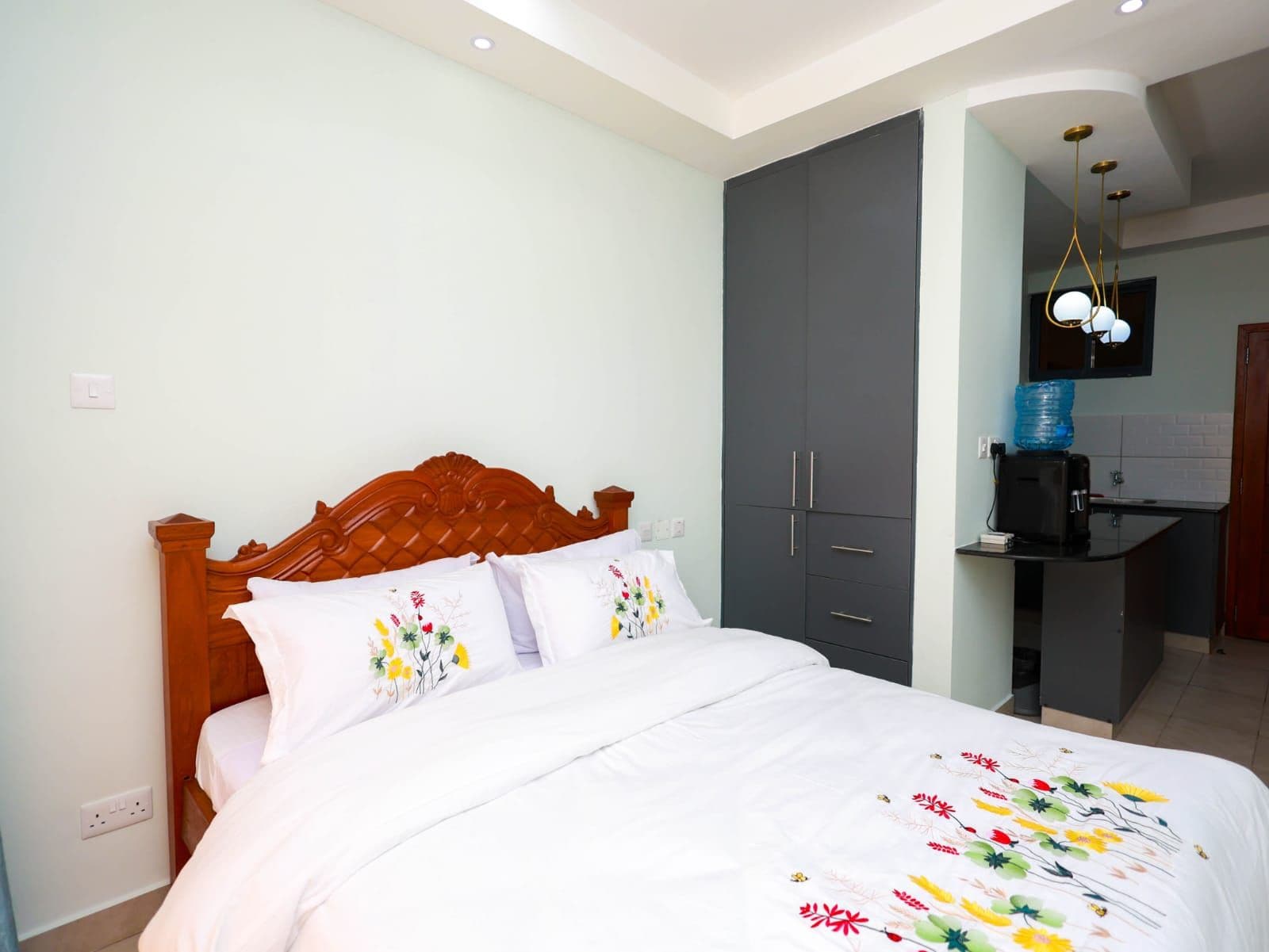 A bedroom with a wooden headboard, a bed with white bedding, a grey wardrobe, a small kitchenette with a coffee machine, and a wall-mounted TV.