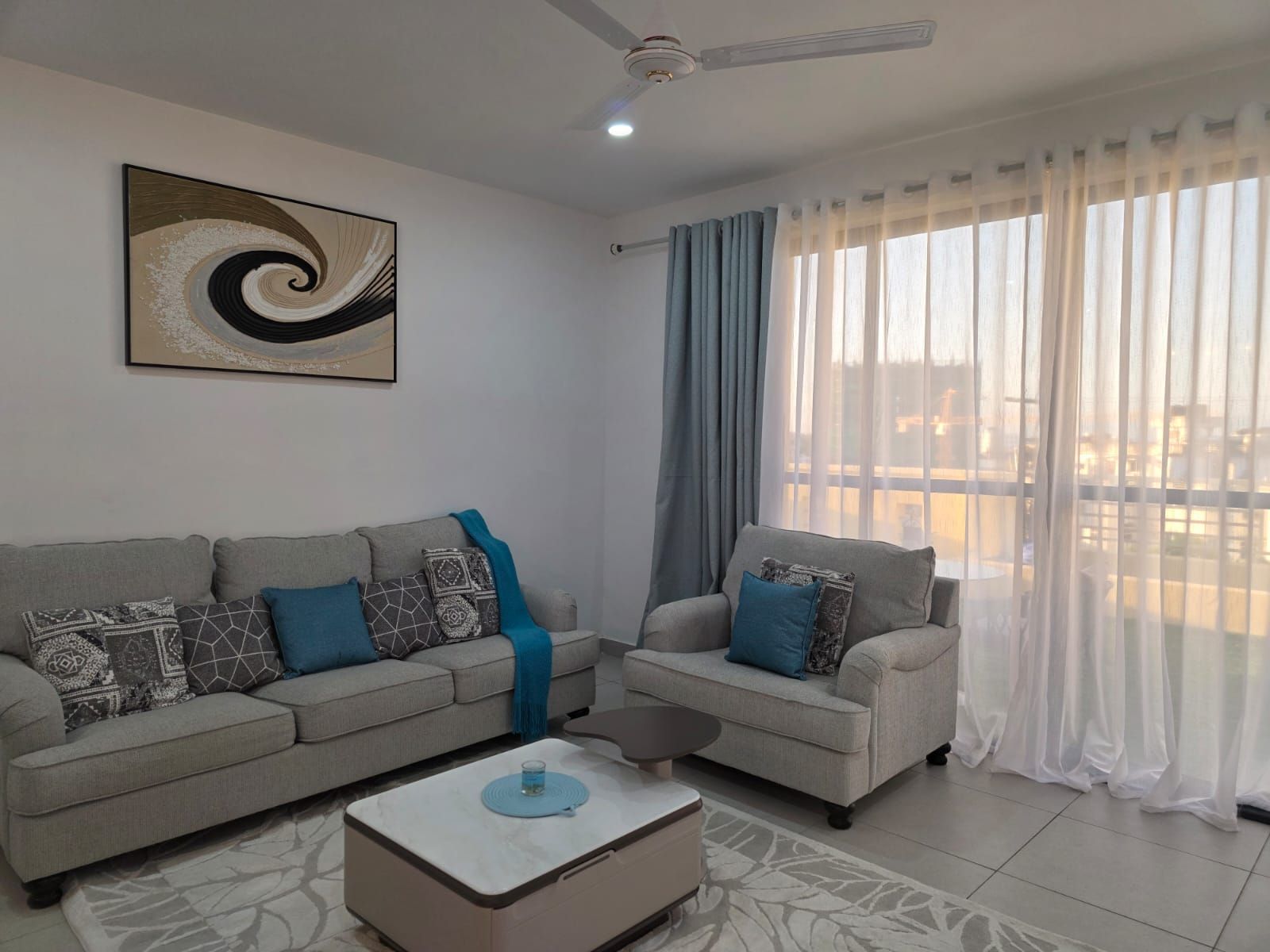 A living room with two sofas, a coffee table, a ceiling fan, and a large window with sheer curtains.