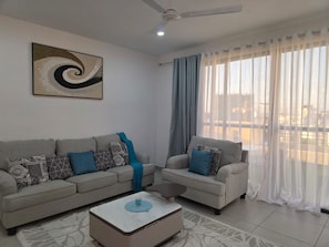 Ein Wohnzimmer mit zwei Sofas, einem Couchtisch, einem Deckenventilator und einem großen Fenster mit durchsichtigen Vorhängen.