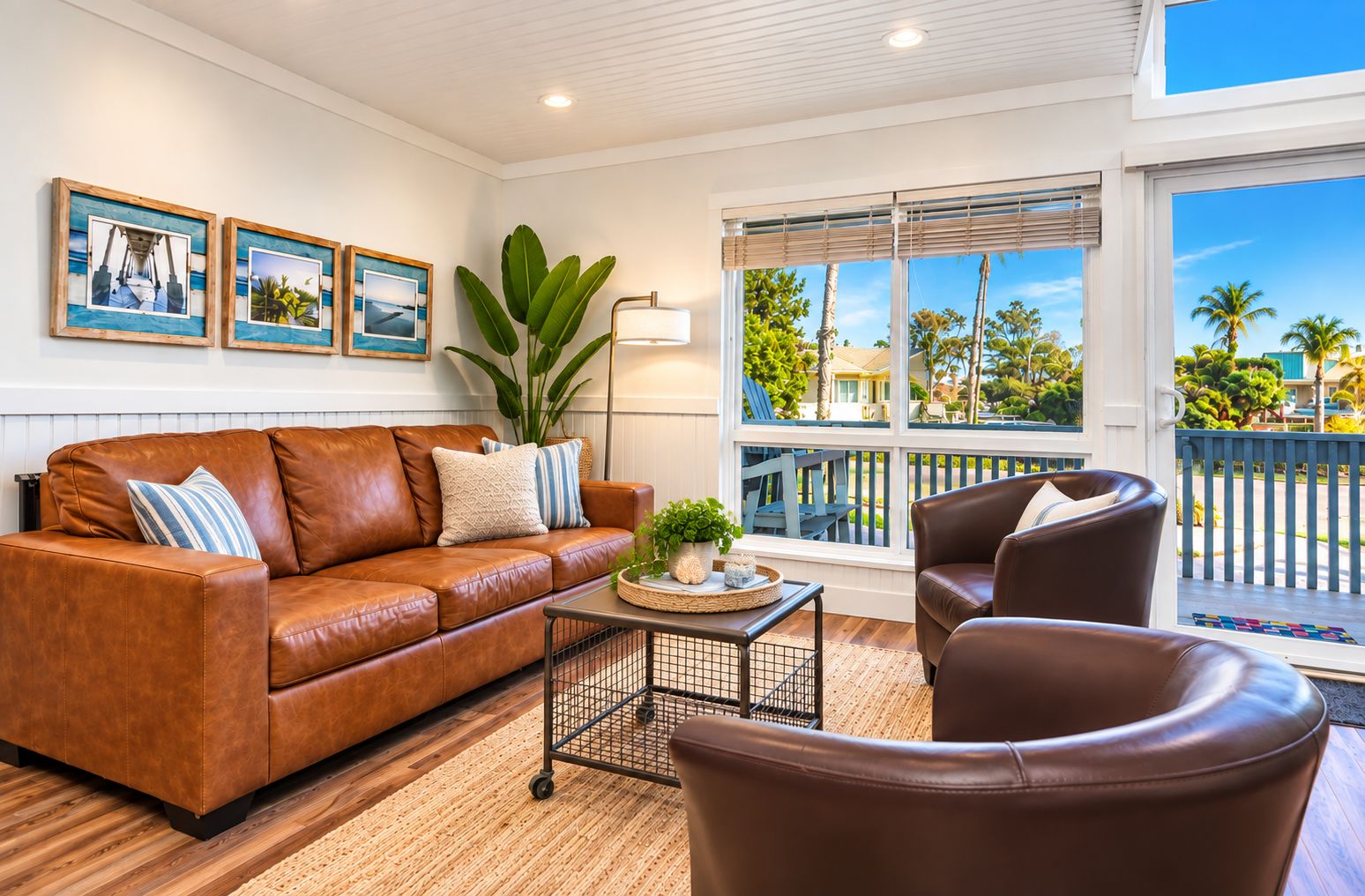A living room with a brown leather sofa, a coffee table, and a large potted plant. There are three framed pictures on the wall, and a window with a view of palm trees.