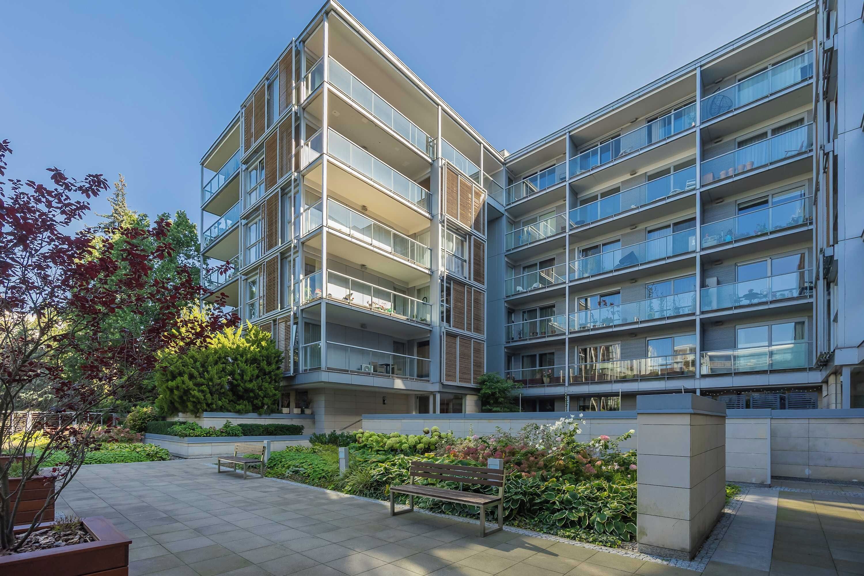 A modern multi-story residential building with balconies, a paved courtyard, and a bench.