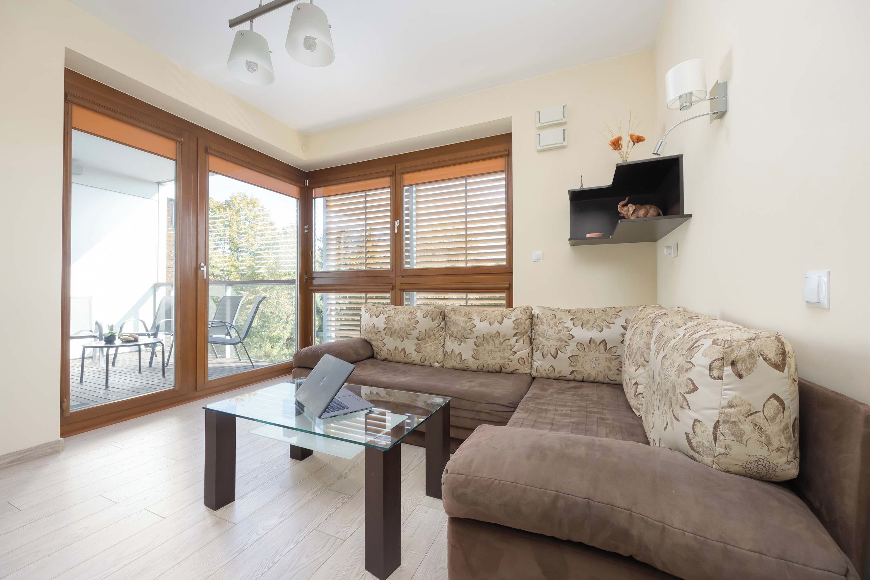A modern living room with a glass coffee table, a brown sofa with floral cushions, and large sliding glass doors leading to an outdoor patio.