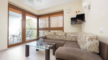 A modern living room with a glass coffee table, a brown sofa with floral cushions, and large sliding glass doors leading to an outdoor patio.
