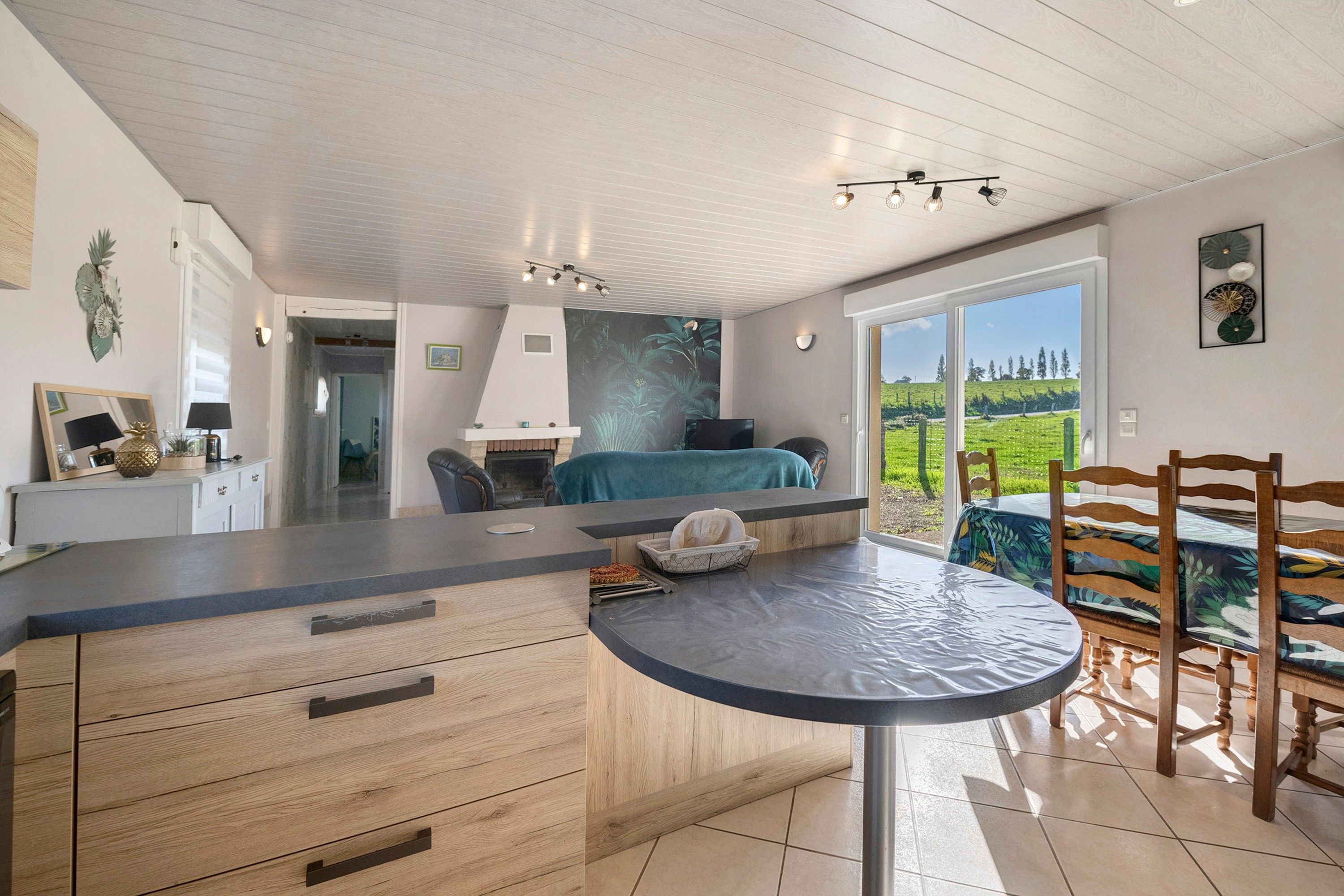 A modern kitchen with a central island, a dining area with a round table, and a view of a green landscape through large windows.