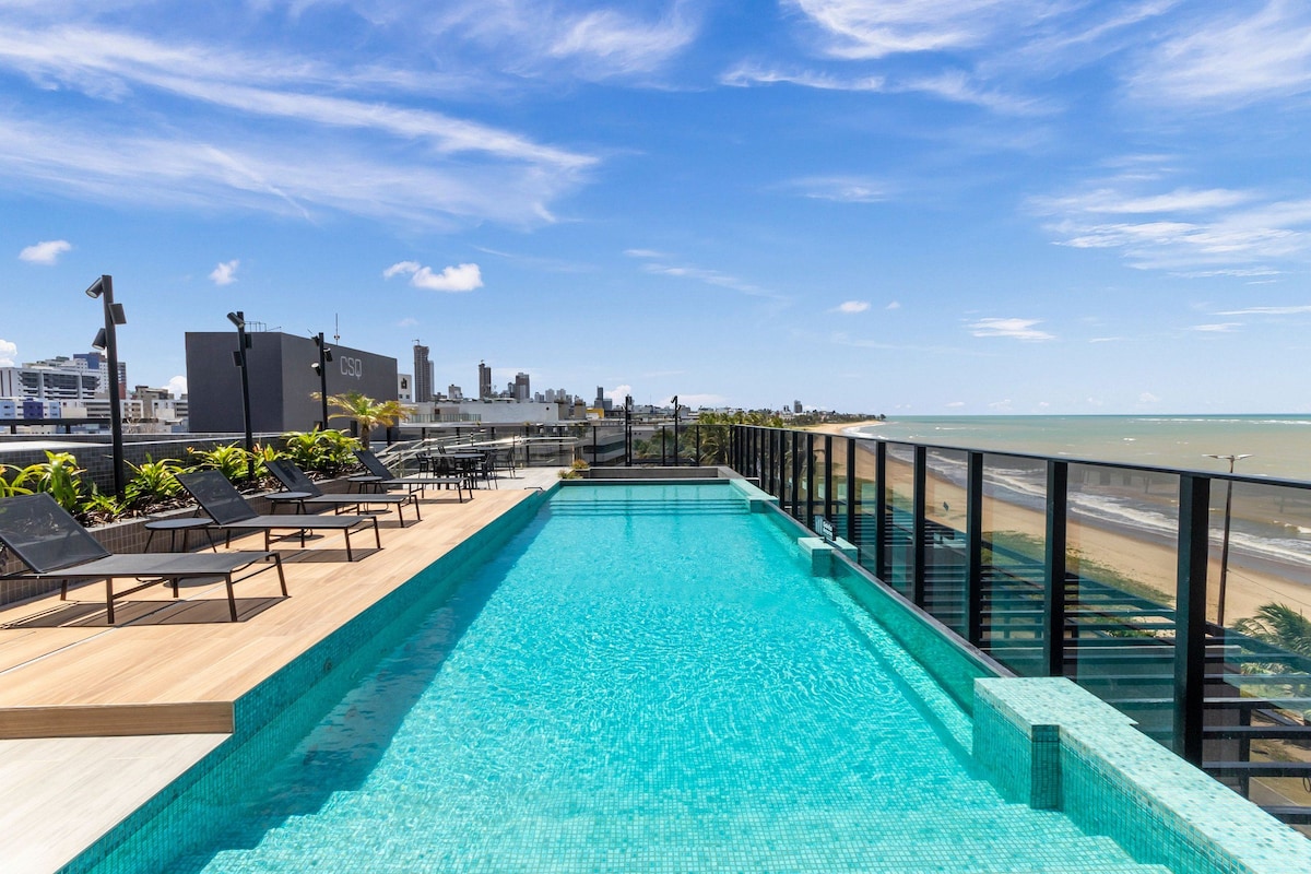 Uma piscina infinita no terraço com vista para a cidade, espreguiçadeiras e guarda-corpo de vidro com vista para a praia.