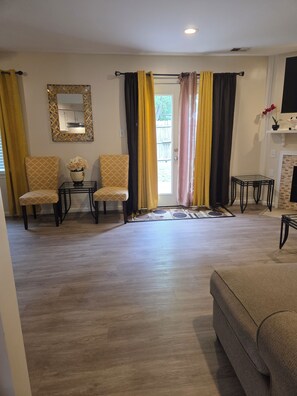 Un salon avec deux fauteuils à motifs, une petite table sur laquelle est posé un vase, un canapé, une cheminée et un miroir au mur.