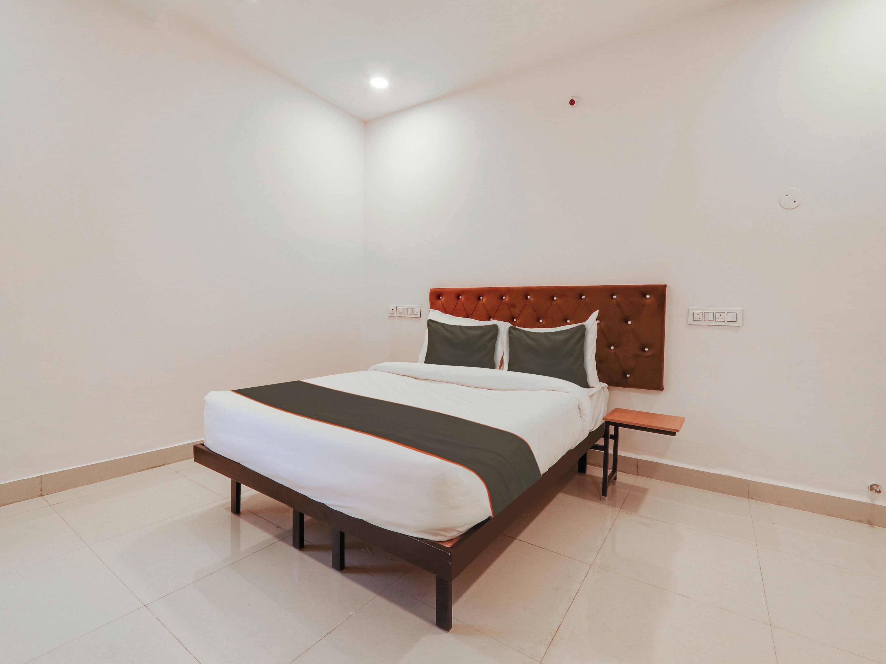 Una cama bien hecha con cabecera marrón, una mesita de noche y un enchufe en la pared.