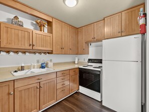 A kitchen with wooden cabinets, a white refrigerator, a stove, and a countertop with various items. - Summit Nook | 2 bdrm Brian Head Condo (Brian Head)
