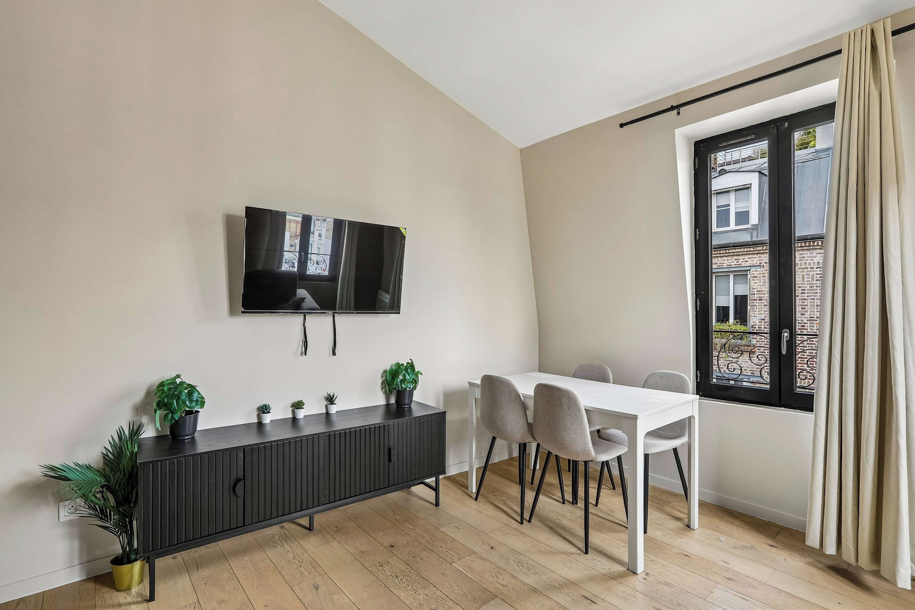 A modern living room with a flat-screen TV mounted on the wall, a dark wooden console table with potted plants, a white dining table with four chairs, and a large window with a view of a building.