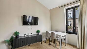A modern living room with a flat-screen TV mounted on the wall, a dark wooden console table with potted plants, a white dining table with four chairs, and a large window with a view of a building.
