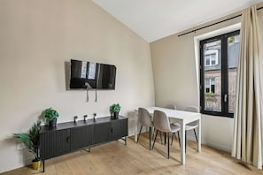 A modern living room with a flat-screen TV mounted on the wall, a dark wooden console table with potted plants, a white dining table with four chairs, and a large window with a view of a building. - Cozy 1BR Retreat for Couples & Families (Boulogne-Billancourt)