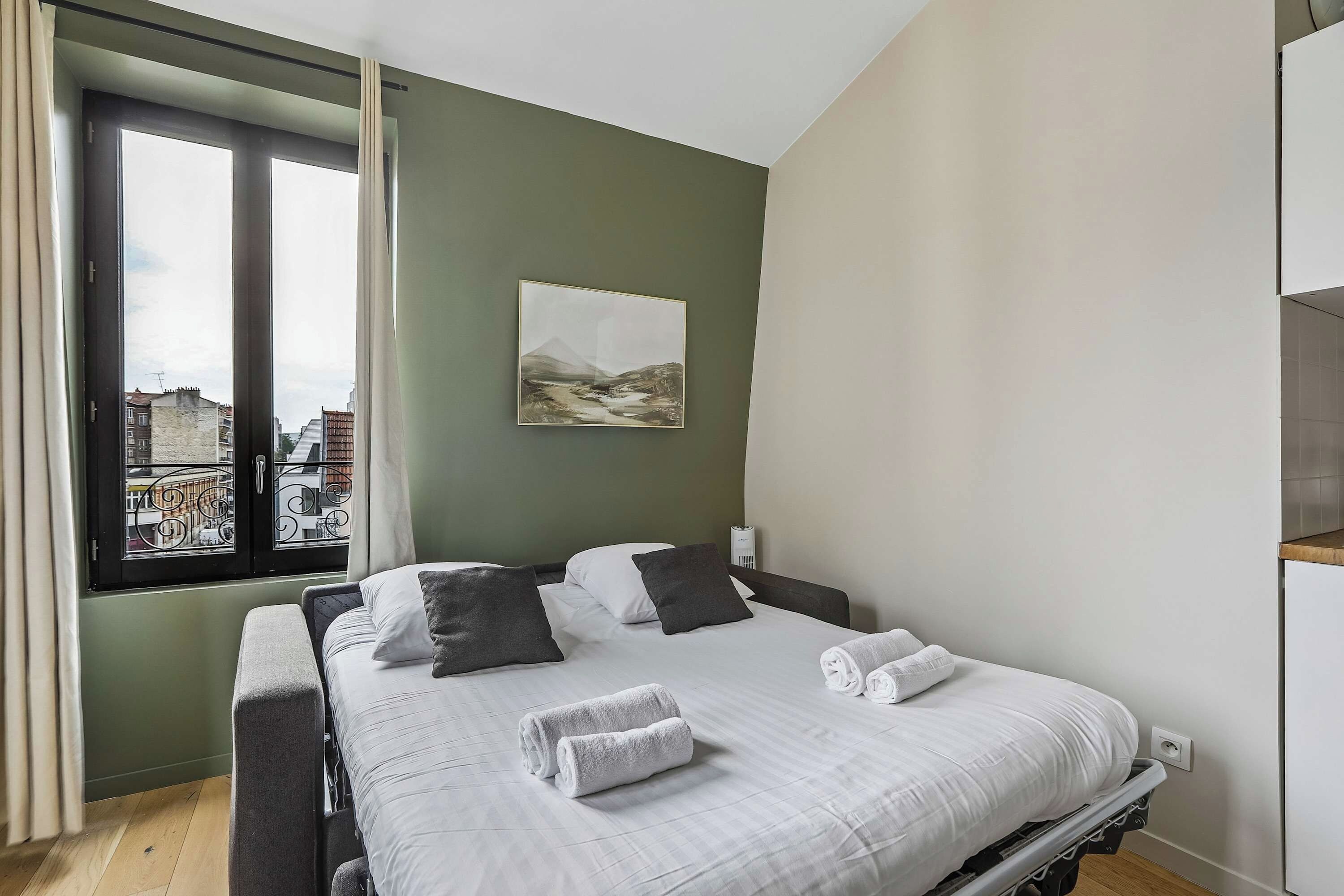 A bedroom with a bed, pillows, a view of buildings through a window, and a painting on the wall.