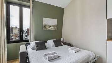A bedroom with a bed, pillows, a view of buildings through a window, and a painting on the wall.