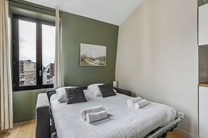 A bedroom with a bed, pillows, a view of buildings through a window, and a painting on the wall. - Cozy 1BR Retreat for Couples & Families (Boulogne-Billancourt)
