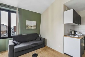 Un apartamento moderno con un sofá gris, una mesita redonda, una cocina con armarios blancos y una ventana con vista a la ciudad.